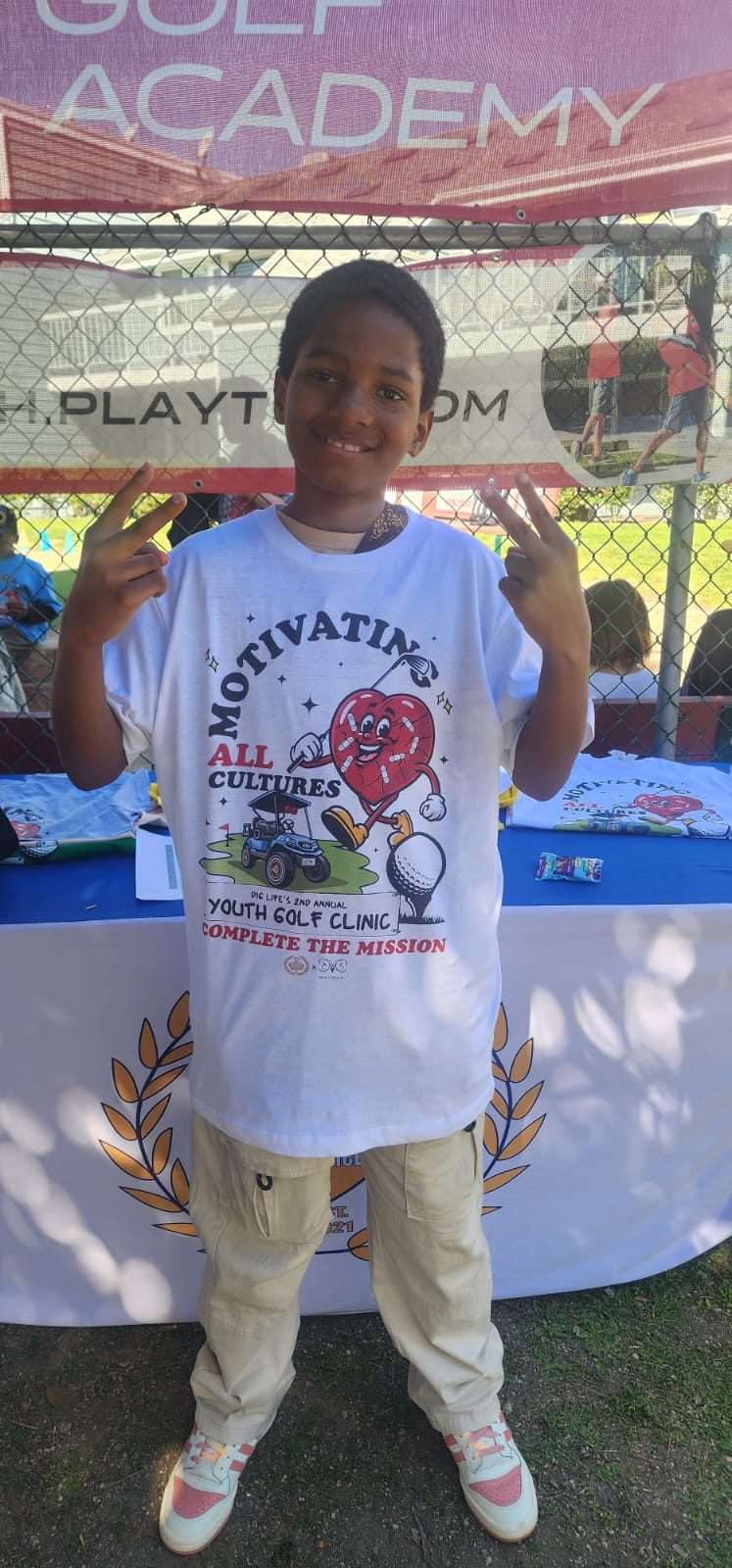A student in a Motivating All Cultures Youth Golf Clinic shirt throwing up peace signs.
